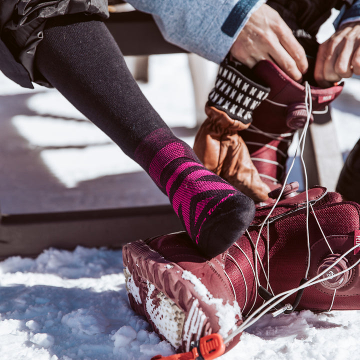 Model in the snow wearing RFL over-the-calf ski & snowboard sock in black while lacing up her snowboard boots