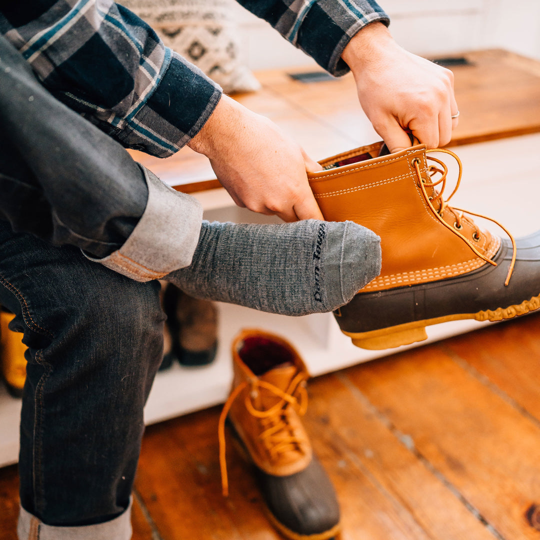 Man sitting on a wooden bench, putting on boots, wearing The Standard Mid-Calf Lightweight Lifestyle Sock in Medium Gray