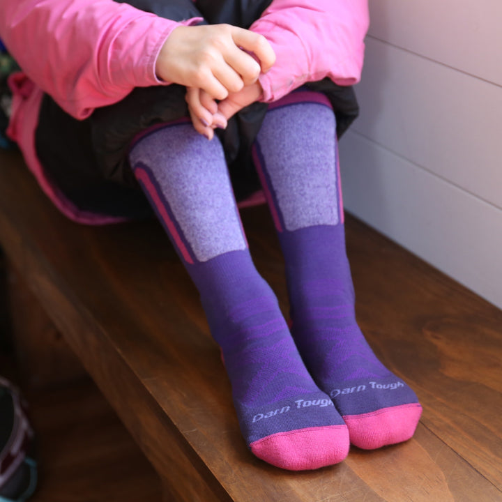 model sitting on a wood bench wearing the 3810 T-Bar over the calf ski and snowboard sock in grape colorway