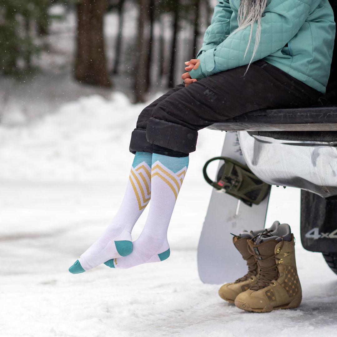 Model sitting in car trunk wearing women's peak over-the-calf snow sock in white with brown boots on ground