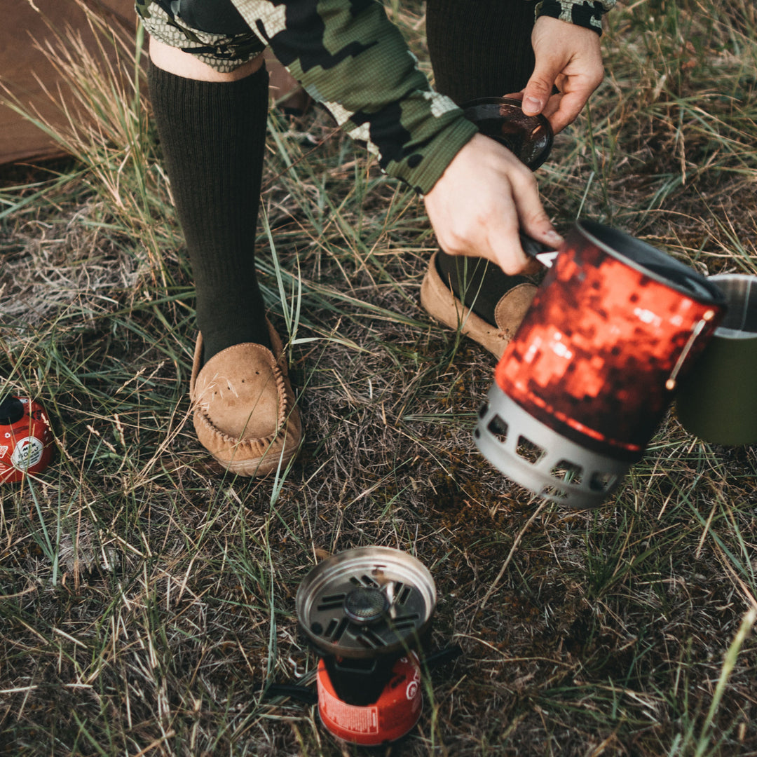 Image of a man wearing slippers at a campsite on the grass, in camo gear pouring coffee out of a thermos while wearing T3005 Mid-Calf Lightweight Tactical sock in Black, Lifestyle Image