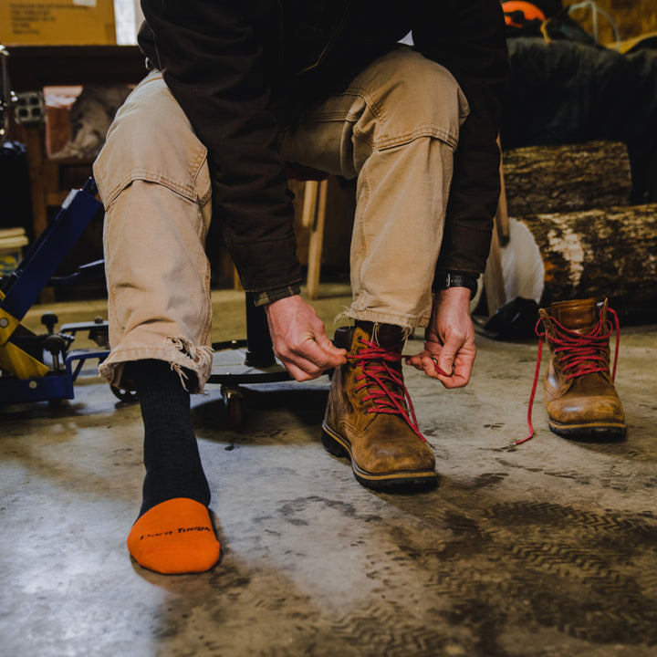 Close up shot of model sitting on a shop stool wearing 2007 socks in graphite colorway and lacing up brown work boot on left foot.