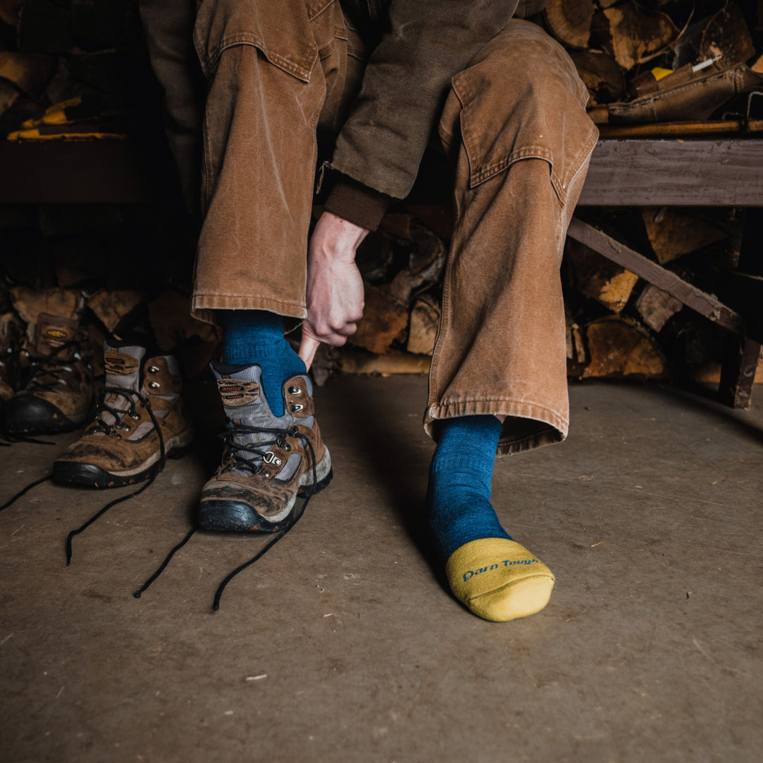 Model sitting on bench in front of wood pile wearing 2016 socks in indigo colorway and pulling on brown work boot on right foot. 