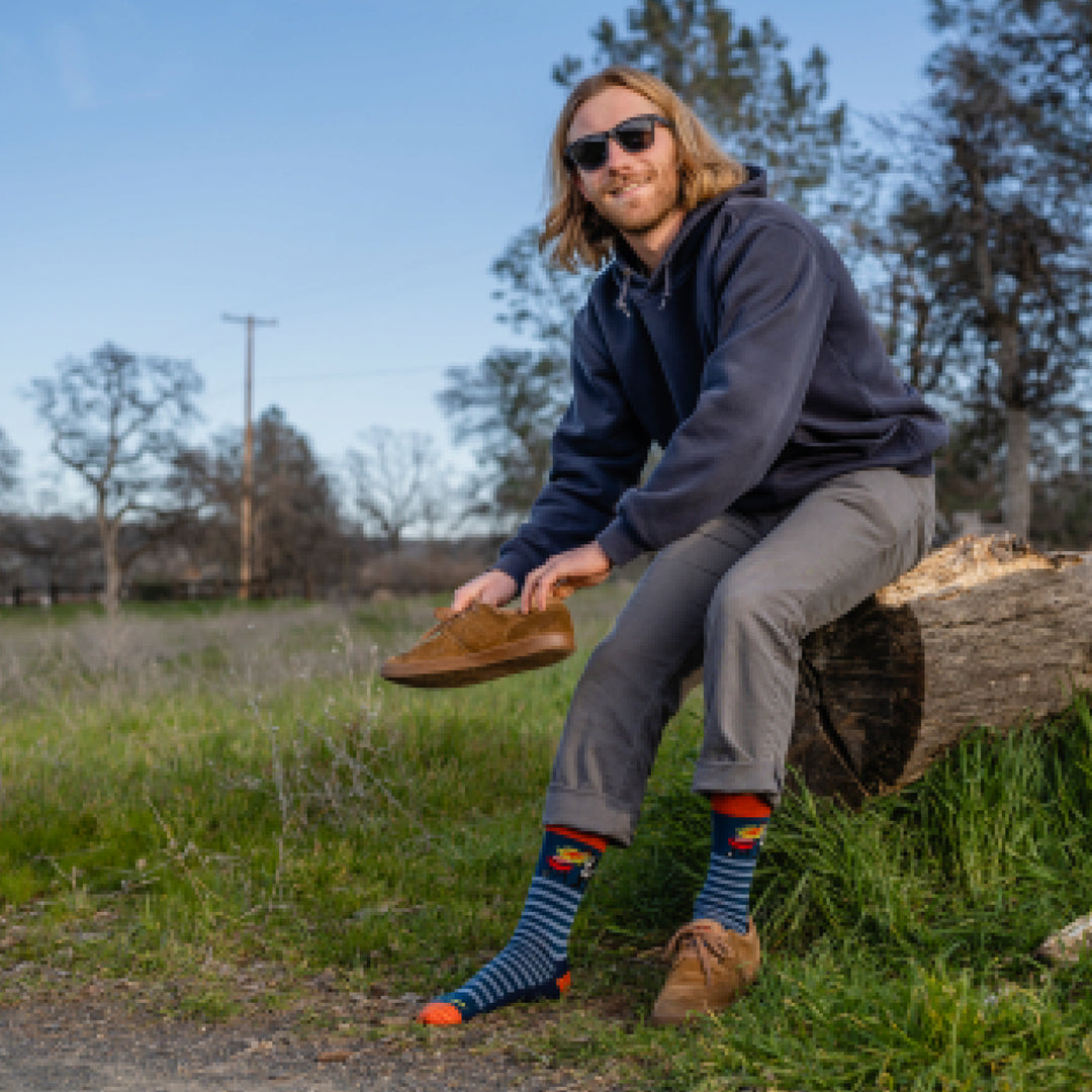 Model sitting on a rock while putting on a brown sneaker and wearing 6066 socks in Deep Water colorway