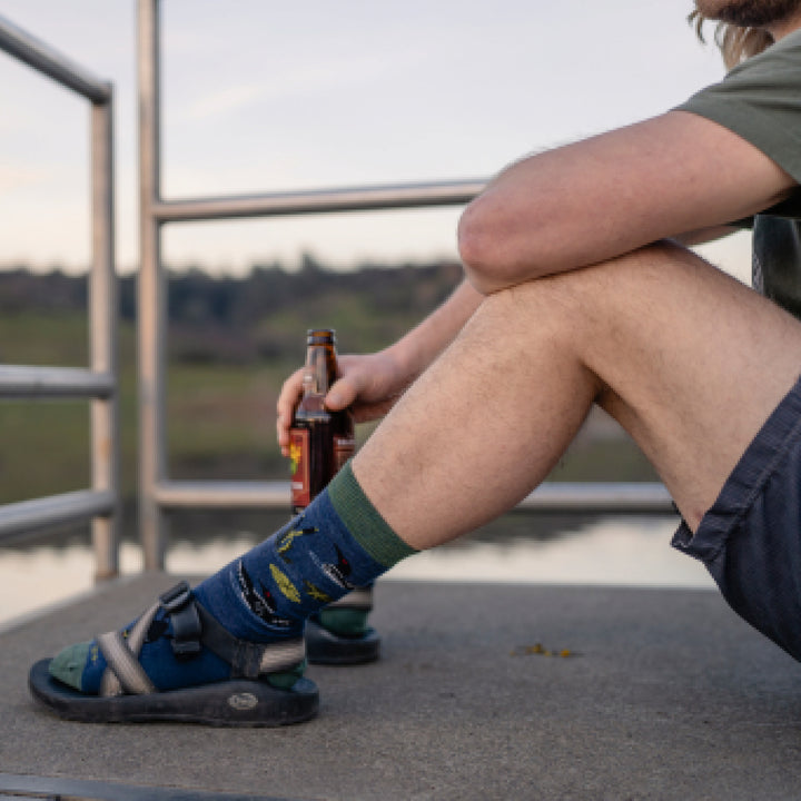 Model sitting on dock holding a bottle of beer and wearing 6112 socks in denim colorway with hiking sandals