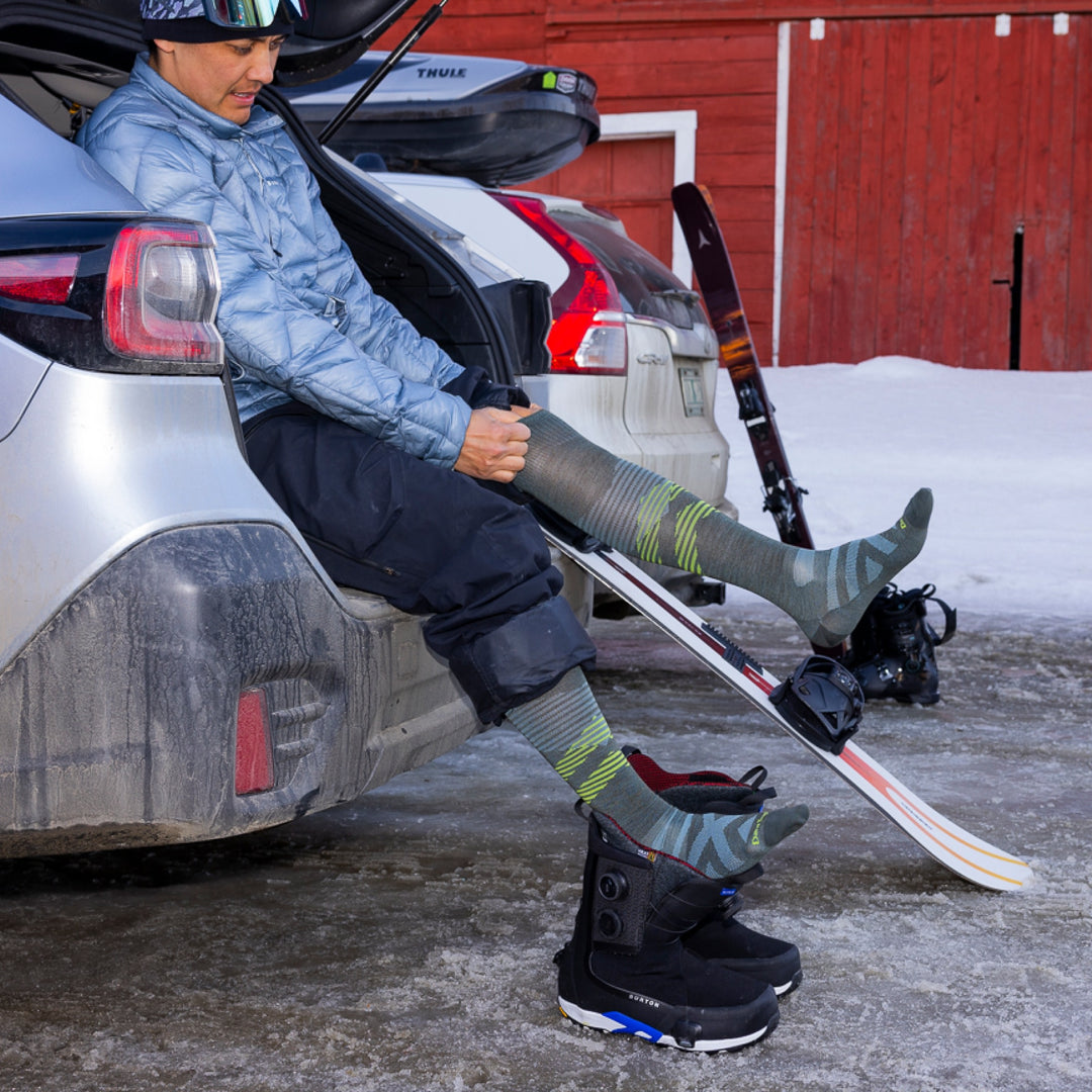 model sitting on edge of car trunk with snowboard and snowboard boots putting on 8002 pennant RFL OTC ultra lightweight sock forest