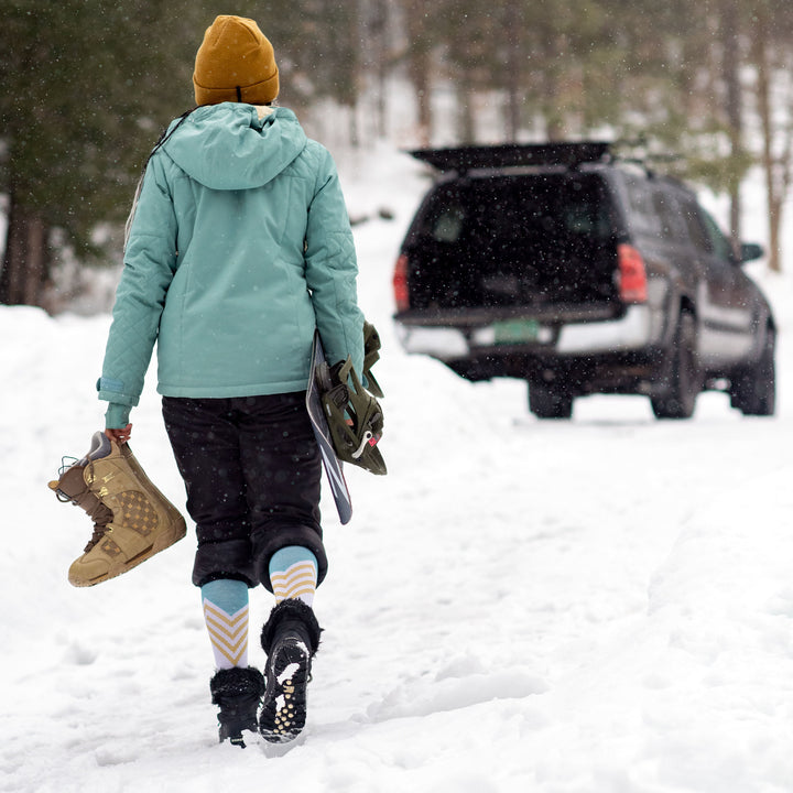 Model walking through snow wearing women's peaks over-the-calf snow sock in white while holding snowboard and boots