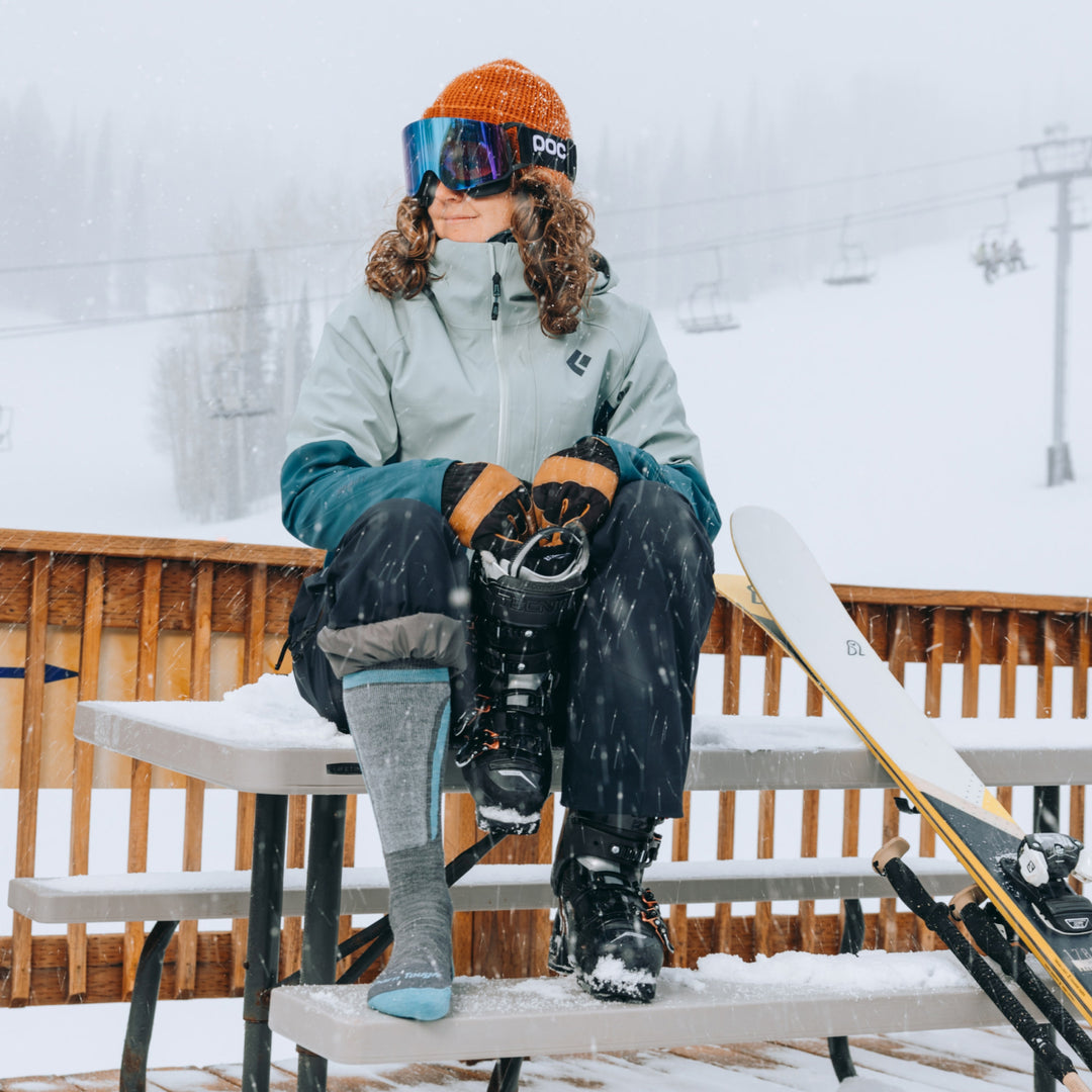 model sitting on a picnic table holding one snow boot wearing the 8057 edge ski and snowboard sock in gray 