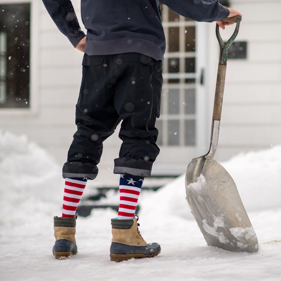 Model shoveling snow wearing men's captain stripe over-the-calf snow sock with gray and brown snow boots