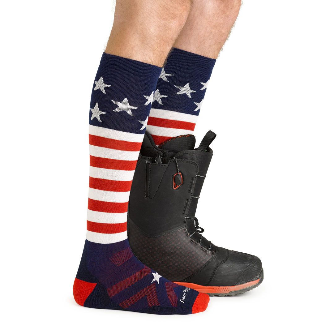 On model side view, front foot barefoot wearing Captain Stripe Over the Calf Ski & Snowboard Socks in Stars & Stripes pattern and back foot wearing a snowboard boot