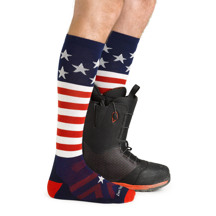On model side view, front foot barefoot wearing Captain Stripe Over the Calf Ski & Snowboard Socks in Stars & Stripes pattern and back foot wearing a snowboard boot