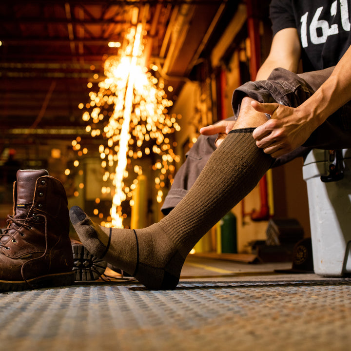 Side shot of model sitting on a cooler in a workshop putting on the men's paul bunyan over-the-calf work sock in timber brown