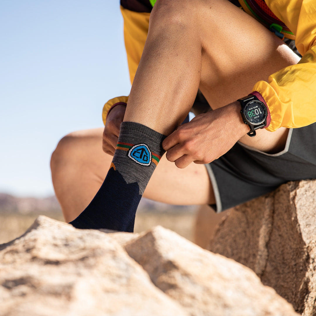 Model sitting down on rocks in Joshua Tree putting on the CDT micro crew hiking sock in eclipse blue