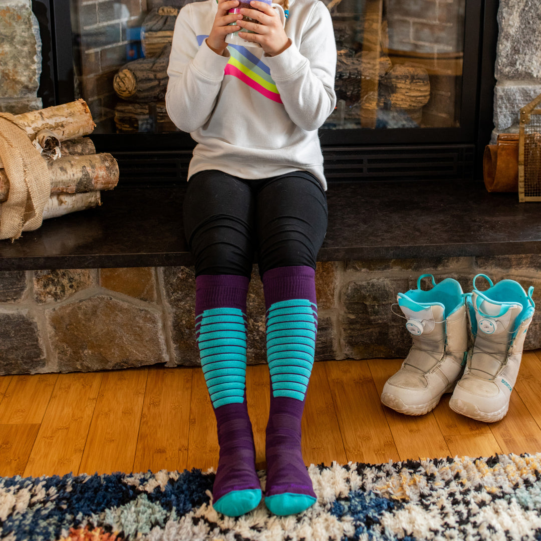 Close up shot of girl wearing Edge Jr snow socks, sitting on the edge of the fireplace drinking hot cocoa.