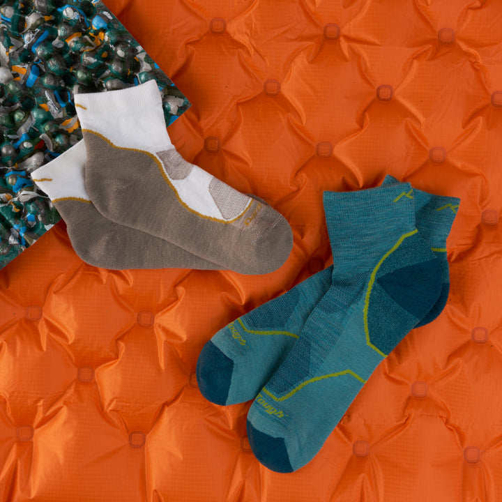 2 pairs of lightweight quarter hikers 1 men's in aqua 1 women's in oat milk laying on orange sleeping bag 