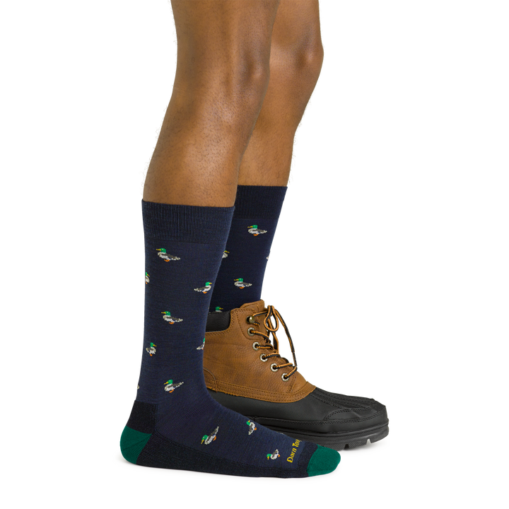 Side studio shot of model wearing men's duck duck moose crew lifestyle sock in navy eclipse with brown boot on left foot