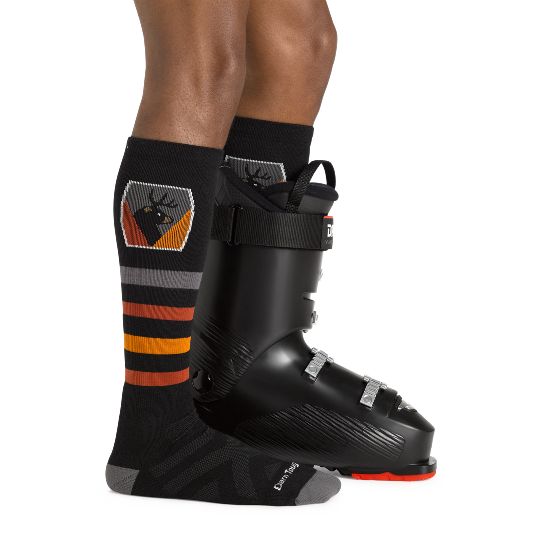 Man facing to the right, wearing Beer Badge Over the Calf Ski & Snowboard Socks in Black, back foot also wearing a ski boot