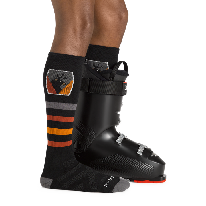 Man facing to the right, wearing Beer Badge Over the Calf Ski & Snowboard Socks in Black, back foot also wearing a ski boot