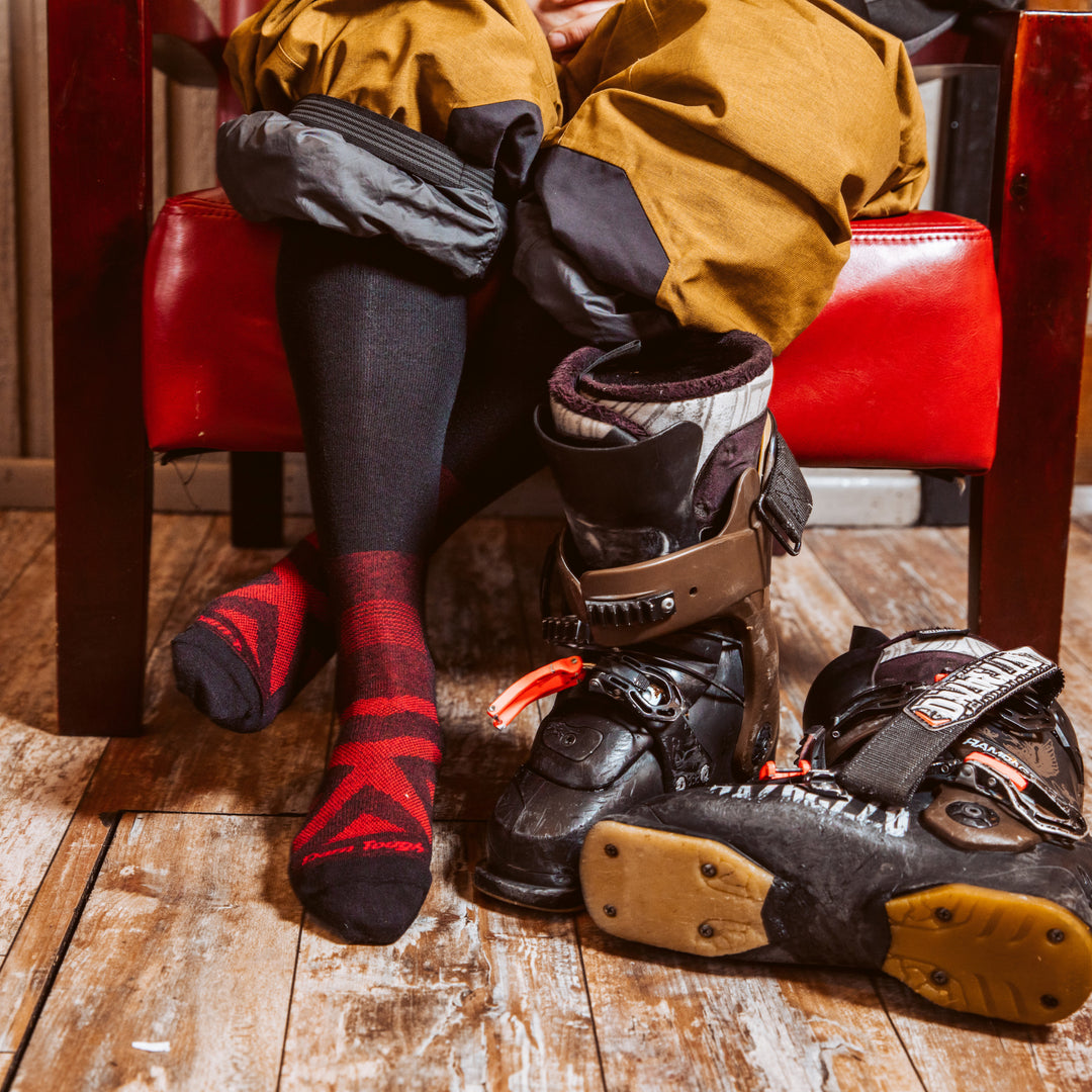 Close up shot of model sitting in a red chair in snowboard gear wearing the men's thermolite over-the-calf snow sock in black