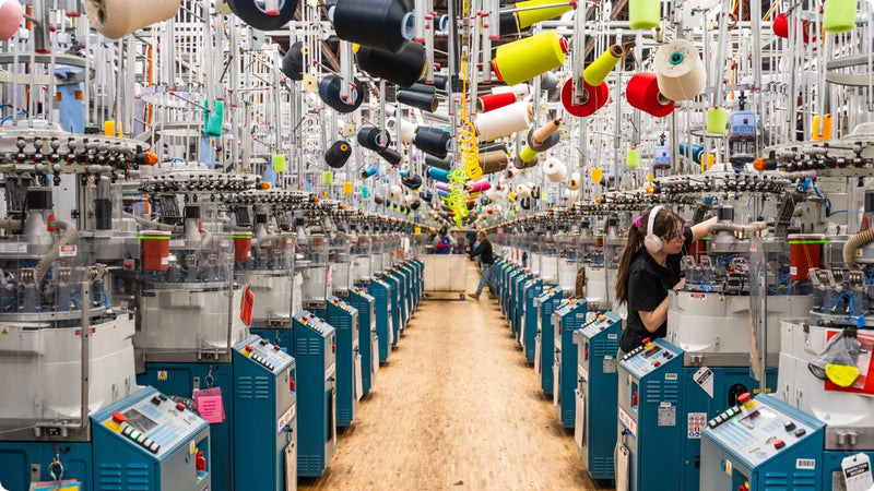 A view of the knitting room floor, full of knitting machines and colorful yarn cones hanging overhead